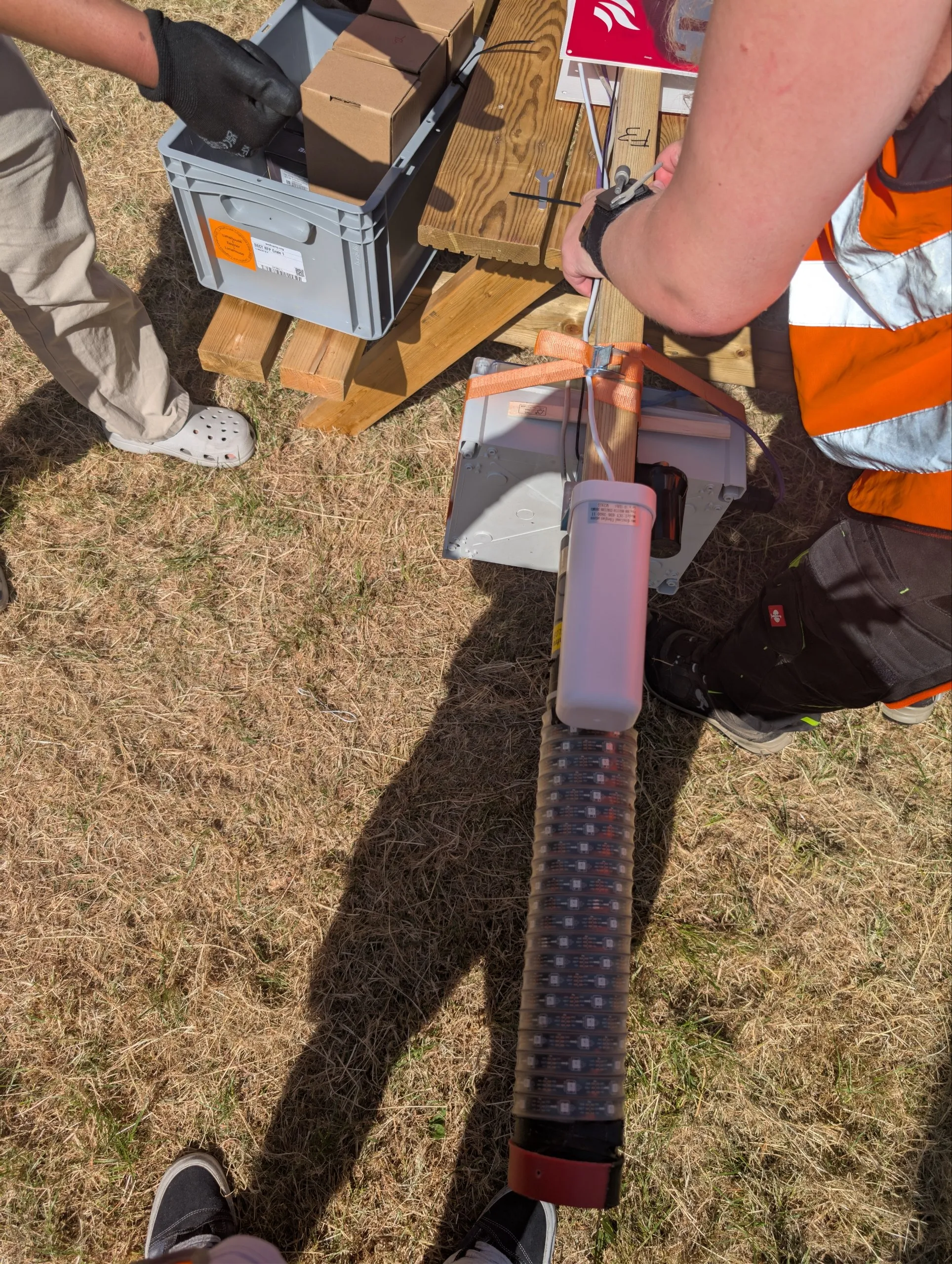 a dk pole being assembled, this one with a weatherproof box holding an access point tied to the pole with a strap, an OHM-light, and an extra 5g antenna.