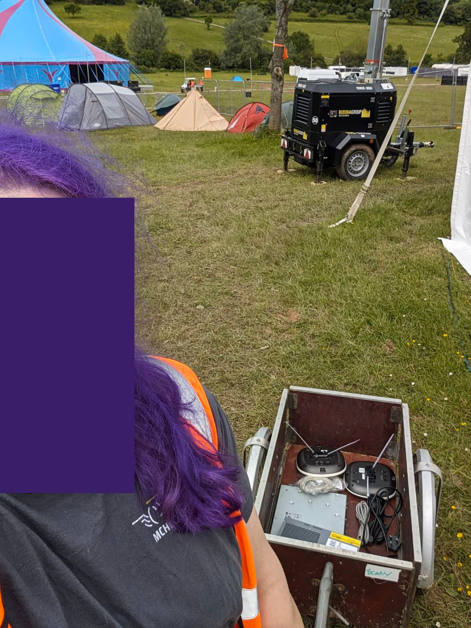 Selfie of myself (obscured) wearing a high-vis jacket, an MCH 2022 t-shirt, and pulling a wooden cart behind me which contains two network switches and two network access points. You can see various tents as well as the event stage in the background.