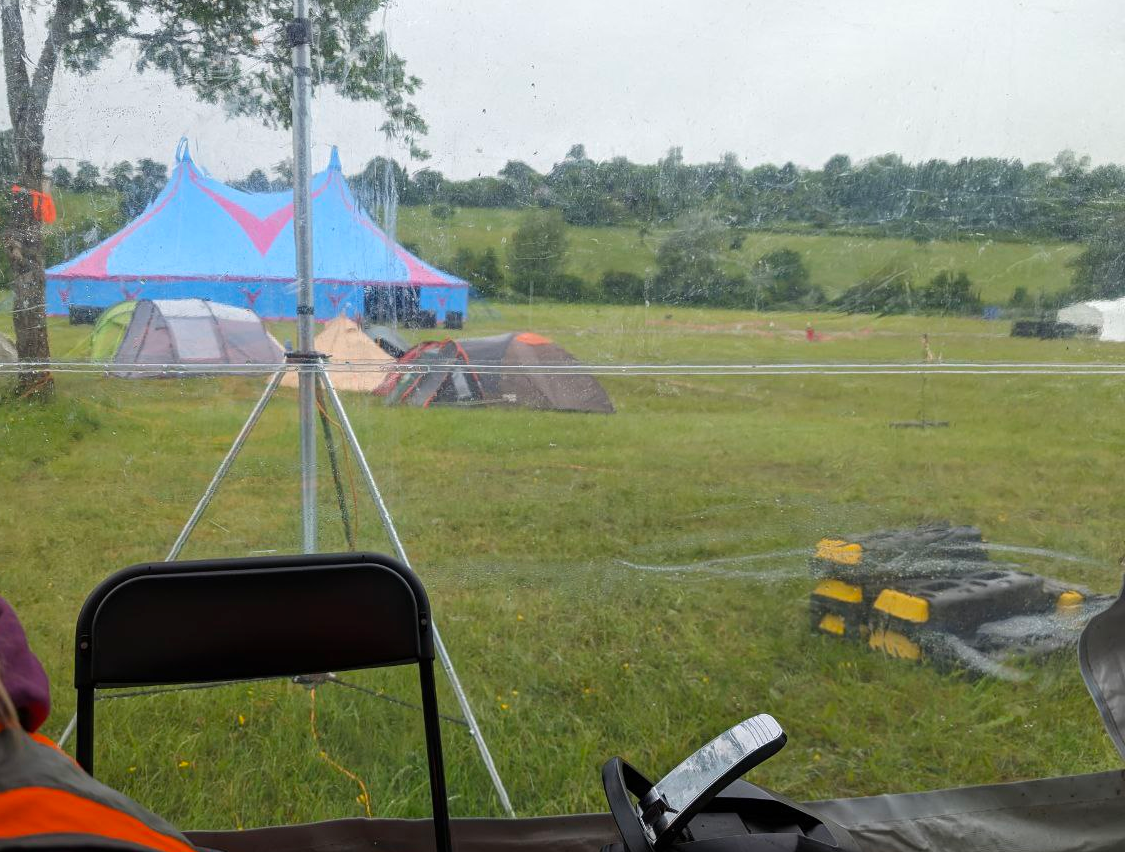 The view from the HQ tent. One can see a few tents next to a tree, an antenna of questionable origin, and one of the event stages in the background.