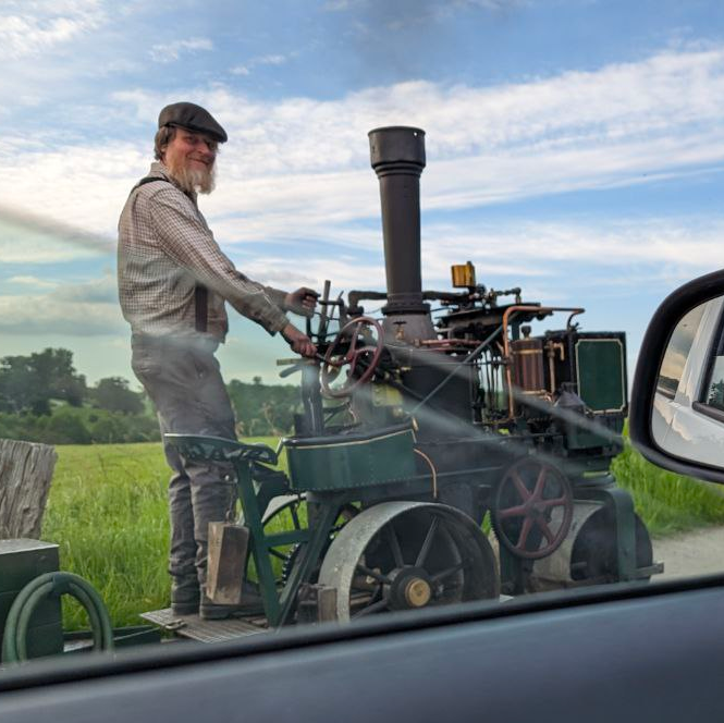 A green steam powered cart, driven by a bearded man in gray overalls.