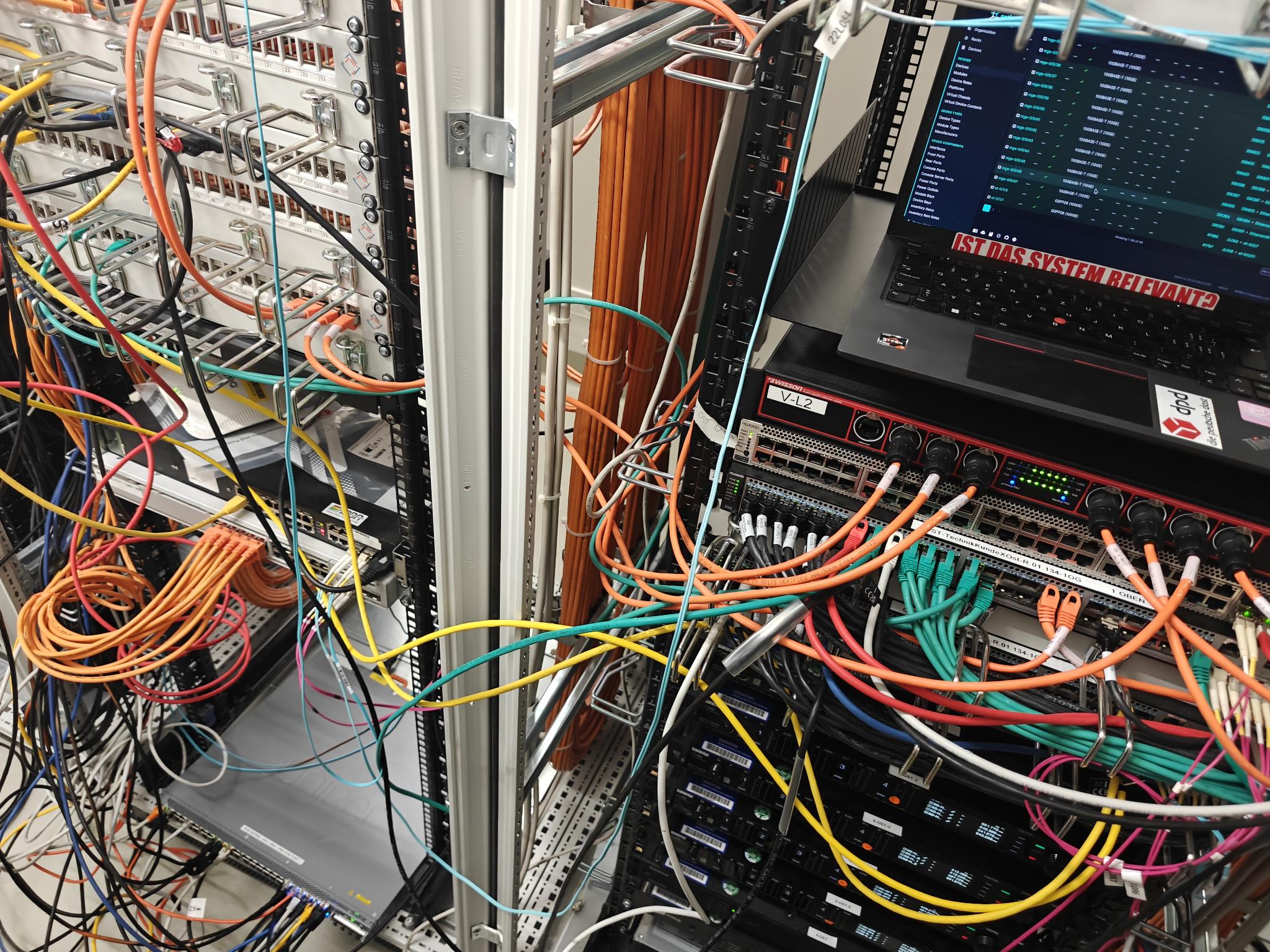 picture of cables in a patch room, spaghetti is starting to form. a laptop with netbox can be seen propped in an empty space in the rack