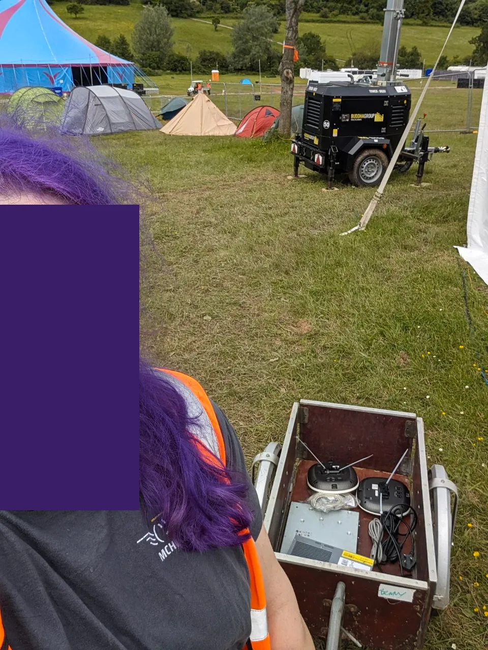 Selfie of myself (obscured) wearing a high-vis jacket, an MCH 2022 t-shirt, and pulling a wooden cart behind me which contains two network switches and two network access points. You can see various tents as well as the event stage in the background
