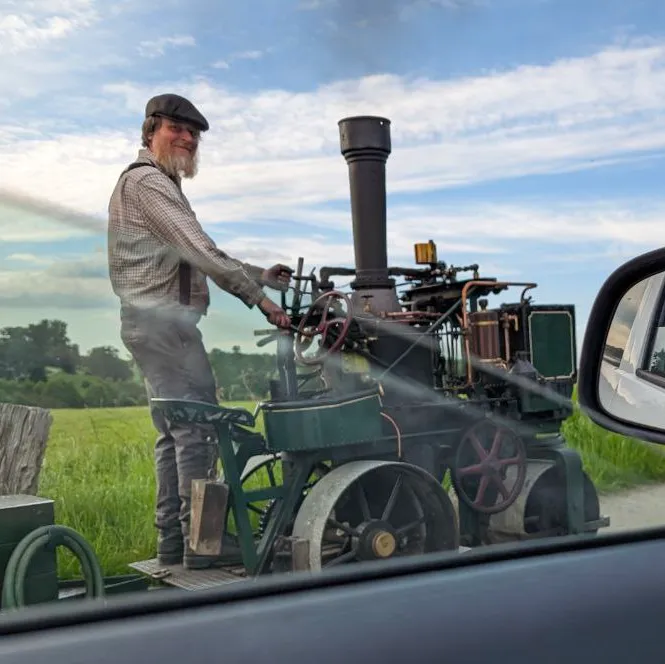 A green steam powered cart, driven by a bearded man in gray overalls